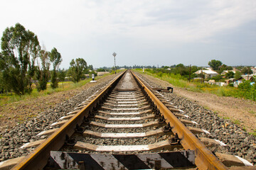 Fototapeta premium Railroad over the horizon. Railway rails and concrete sleepers against the backdrop of a rural area with one-story houses, trees and a blue sky with clouds, goes into the distance. Travel concept