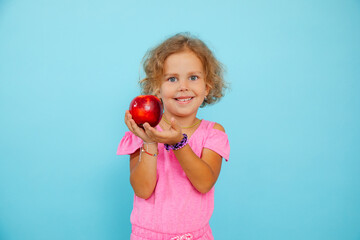 Portrait of little girl with curly fair hair wearing pink jumpsuit, holding showing big red apple on blue background.