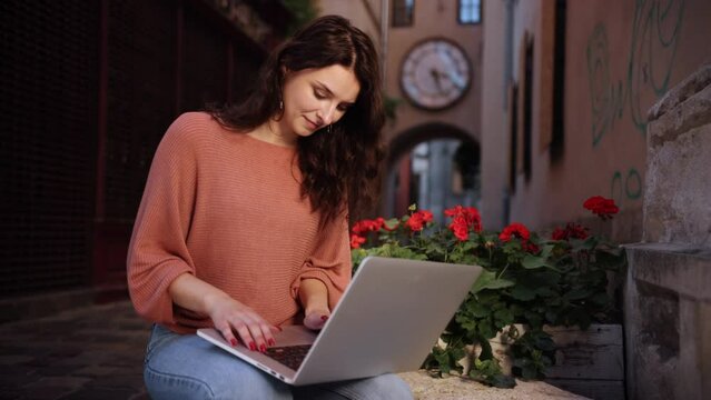 Pretty female student studying outdoors in nature, attractive female using laptop while sitting on decorative stone of city streets, adult lady studying outside in nature on a laptop keyboard