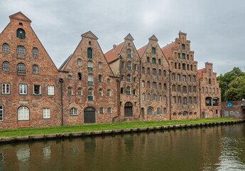 Fototapeta na wymiar Germany, Lubeck - July 13, 2022: Row of red-brown historic salt storage warehouses along Trave river under blueish cloudscape. Green lawn on quay.