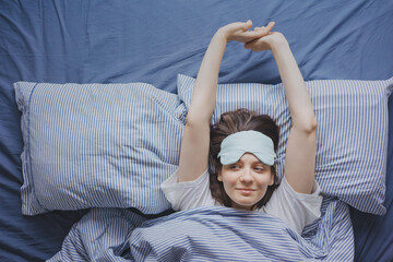 top view of a woman in bed wearing a sleep mask, a happy European woman wakes up and stretches,...