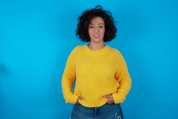 Portrait of successful young beautiful woman with curly short hair wearing yellow sweater over blue...