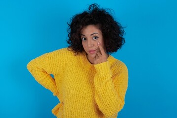 young beautiful woman with curly short hair wearing yellow sweater over blue wall, looking, observing, keeping an eye on an object in front, or watching out for something.