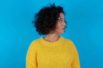 Side view of young happy smiling young beautiful woman with curly short hair wearing yellow sweater over blue wall.