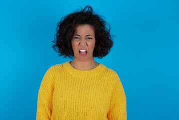 Body language. Disgusted stressed out young beautiful woman with curly short hair wearing yellow sweater over blue wall, frowning face, demonstrating aversion to something.