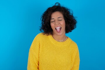 young beautiful woman with curly short hair wearing yellow sweater over blue wall with happy and funny face smiling and showing tongue.