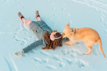 Long curly young girl in pink winter earmuffs and white mittens plays with her dog on winter cold ice river covered with snow. Snowing winter concept.