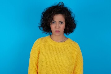 young beautiful woman with curly short hair wearing yellow sweater over blue wall frowning his eyebrows being displeased with something.