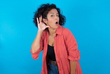 Oh my God! Funny astonished young beautiful woman with curly short hair wearing red overshirt over blue wall opening mouth widely looking aside, with hand near ear trying to listen to gossips.
