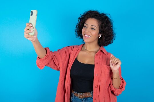 Portrait of a young beautiful woman with curly short hair wearing red overshirt over blue wall  taking a selfie to send it to friends and followers or post it on his social media.