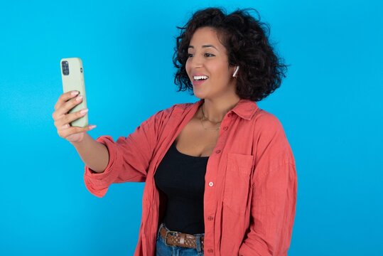 Isolated shot of pleased cheerful young beautiful woman with curly short hair wearing red overshirt over blue wall, makes selfie with mobile phone. People, technology and leisure concept