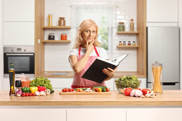 Housewife wearing an apron behind a kitchen counter with vegetables and holding a cook book