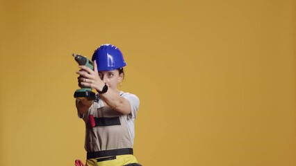 Construction worker having fun with power drill machine, fooling around in studio and playing with electric drilling nail gun over yellow background. Playful female handywoman with screw gun. - Powered by Adobe