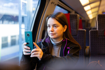 Interested female traveler sitting near window on comfortable seat in express train, filming picturesque mountain landscapes of Switzerland behind glass