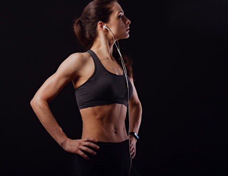 Profile shot of serious sporty female with ponytail, listening music in headphones in sportsbra, standing isolated on black background with empty copy space.