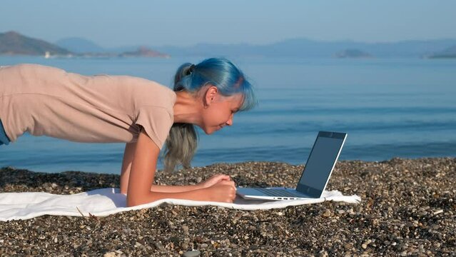 Relaxation exercises online on beach. A teen relax in yoga position against her notebook by the sea under sun.