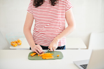 Woman cutting carrots in a kitchen; laptop at the right.