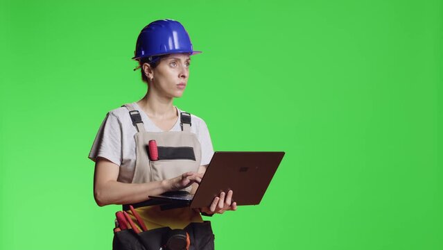 Female construction worker browsing internet on laptop to find refurbishment inspiration, standing over greenscreen isolated background. Woman using pc over chroma key template.