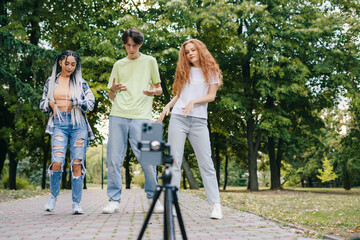 Fototapeta na wymiar Group of trendy friends filming a vlog with a smartphone mounted on a tripod. Having fun dancing in the city park. Listening to music, no stress.