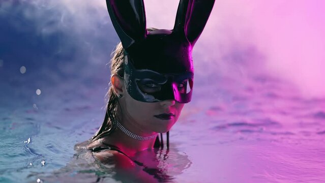 Sexy woman in bunny latex mask swimming in pool at night. Attractive chick in black swimsuit enjoying party time. Rich lifestyle, luxury life, entertainment, masquerade. Dangerous girl