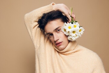 Obraz na płótnie Canvas Close-up photo.A handsome young man with fair skin with dark, short, beautiful hair combed back in a beige turtleneck with a high neck stands on a dark beige background with a bouquet of daisies
