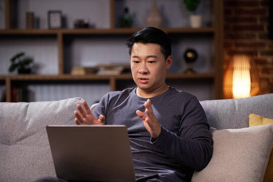 Asian man holding video conference looking at laptop webcam, working from home. Man having online meeting and communicating remotely, sitting on the couch in living room