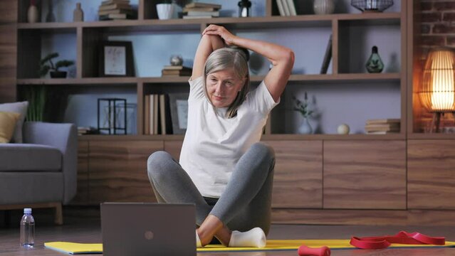 Gray-haired happy mature woman doing gymnastics and yoga at home online with laptop. Active and healthy lifestyle for pensioners. Fitness lessons online, recreation, well-being of the elderly.