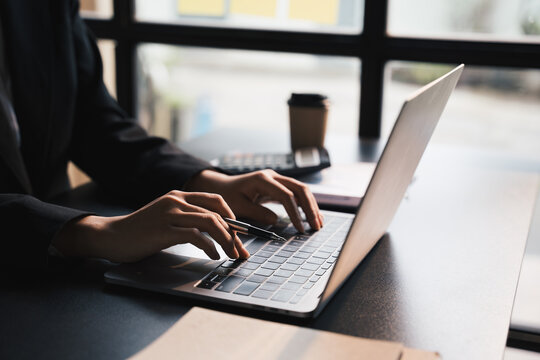 Close-up businessperson typing and searching data internet on laptop and hands holding pen at office desk, Online learning concept.