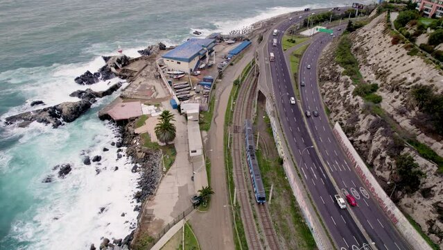 vista aerea desde dron de una zona costera, en donde se ve el mar agitado con olas, una carretera con tr&aacute;fico de veh&iacute;culos y un tren de color azul recorriendo el borde costero