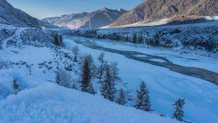 An ice-free turquoise river flows in a snow-covered valley. Spruce trees grow on the banks. A picturesque mountain range against the blue sky is illuminated by the sun. A snowdrift in the foreground. 