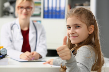 Beautiful little girl shows thumbs up. and doctor sits in background