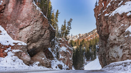 Fototapeta na wymiar The highway runs between sheer cliffs. Coniferous trees grow on the red-brown rocky slopes, there is snow. Blue sky. Red Gate Rocks. Altai
