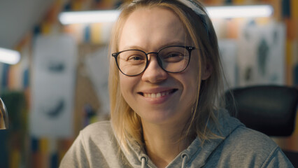 Details of a young blonde woman with a black framed glasses looks at the camera and smiles. Woman sits on a chair in a home based office setup.