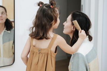 Pretty little girl brushing mother's hair in front of a mirror. Her mom is surprised