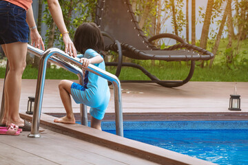 Naklejka premium Adorable daughter in blue swimming suit going down swimming pool ladder to swim with clear blue water and her mother to take care of closely. Little girl at pool. Happy family relationship at pool.