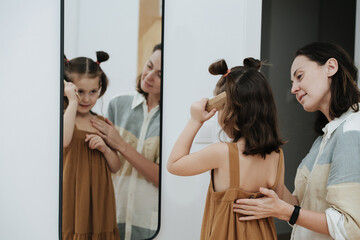 Little girl doing her hair in front of a mirror. Her mom is watching.