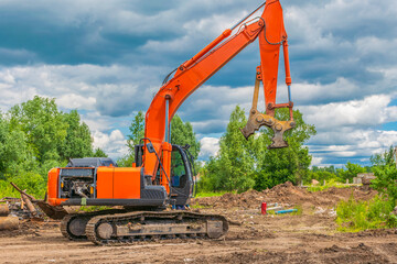 Large orange crawler excavator with crusher rig stands in front of agricultural field with trees and blue cloudy summer sky.