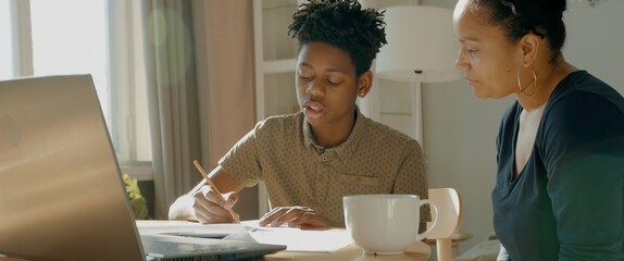 African American Black mother sits near her son, helping him with homework. Distance learning from home during COVID-19 coronavirus pandemic, stay home concept. Shot with 2x anamorphic lens
