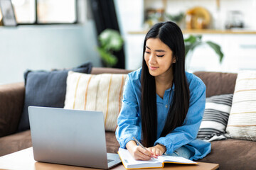 Woman student studying remotely using a laptop, taking notes on notepad sitting from home during online lesson, e-learning concept
