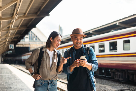 Couple Young asian traveler tourist by train Choose a trip and booking train queue by smartphone. Two Backpacker at platform train station. Happy traveller at railway on vacation holiday weekend.