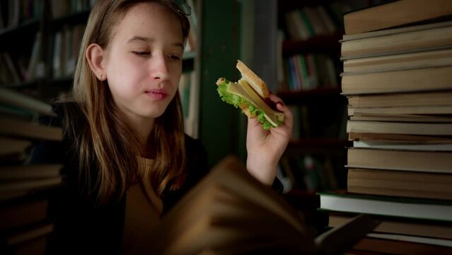Teenager girl sits in library, reads book and eats sandwich. Student receives new knowledge. Smart and well-read person. Education and development. Interesting school literature. Leisure activity.
