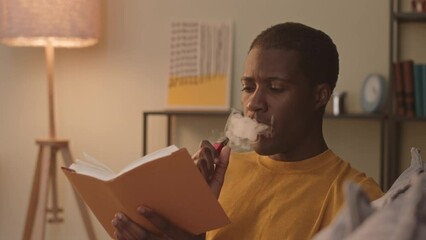 Chest up of young Black man smoking electronic cigarette while reading exciting book at home in evening