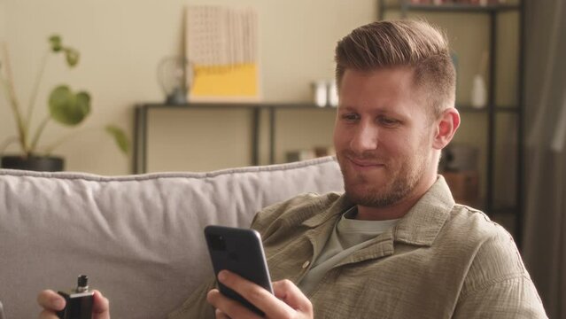 Waist up of young Caucasian man vaping while scrolling feed on smartphone and smiling, siting relaxedly on sofa in living room