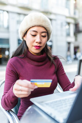 Focused Chinese female with credit card making online purchase on netbook while sitting at table on terrace of outdoor cafe