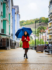 brunette woman in red coat with red lips and blue umbrella walking in the rain on the autumn Kyiv street 