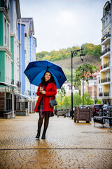 brunette woman in red coat with red lips and blue umbrella walking in the rain on the autumn Kyiv street 
