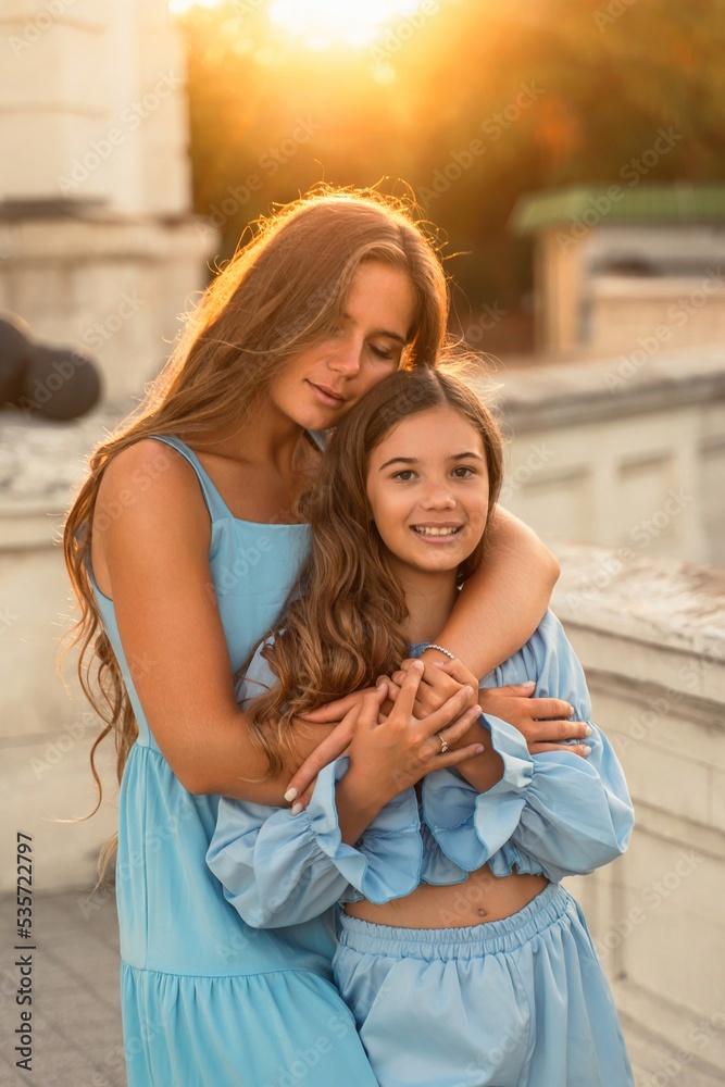 Wall mural Portrait of mother and daughter in blue dresses with flowing long hair against the backdrop of sunset. The woman hugs and presses the girl to her. They are looking at the camera.