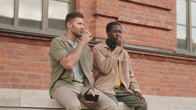 Slowmo of two diverse male friends smoking electronic devices and having conversation outdoors