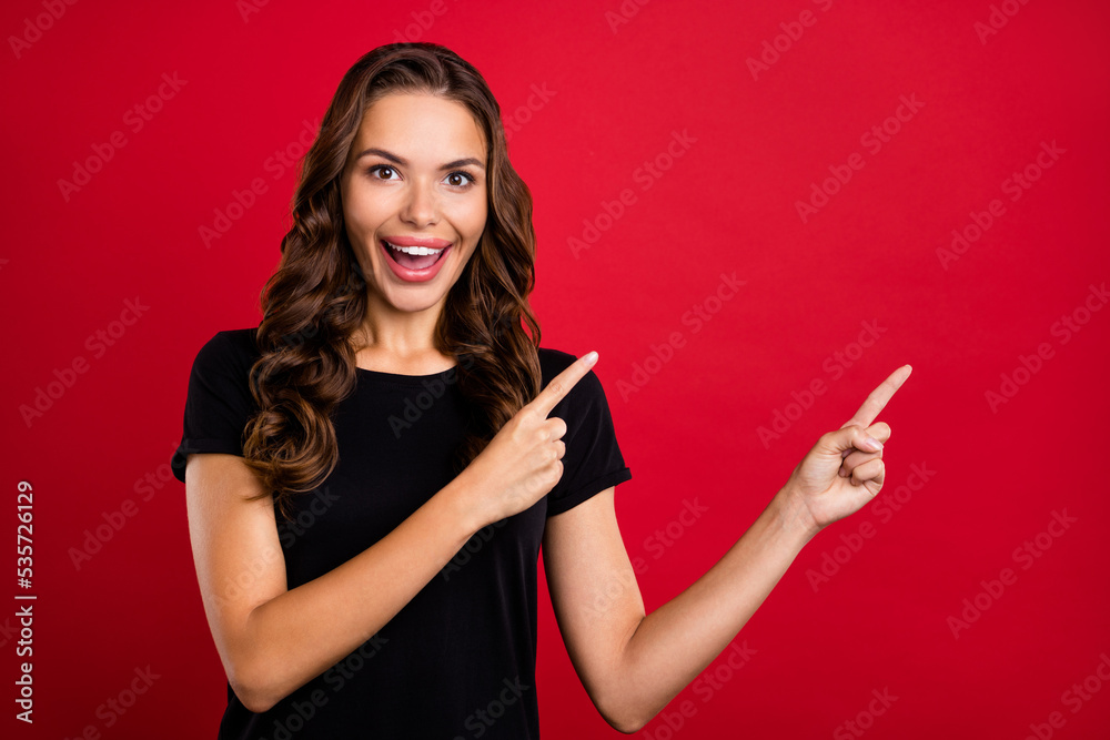 Wall mural Photo of excited pretty young woman wear black t-shirt pointing two fingers empty space smiling isolated red color background