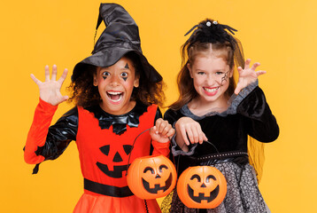 Two happy   children girls in witch  costume and terrible makeup with  pumpkin basket  make scary  gestures and celebrates Halloween and laughs on yellow background